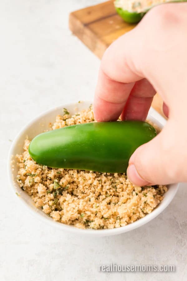 filled jalapeno halves being dipped into breadcrumb topping in a bowl