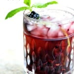 A Spiced Blackberry Cocktail served in a Old fashioned glass garnished with a mint mint leave and a black berry on the rocks