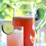Raspberry Limeade displayed in a tall no stem champagne flute garnished with a lime slice, pitcher of Limeade behind champagne flute