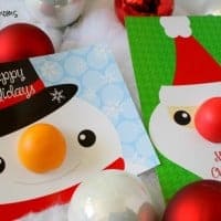 chapstick Holiday Cards. two cards one of snowmans face on a blue card. with EOS orange chapstick as his nose Card says "HAPPY HOLIDAYS" second card santa face on a green card. with EOS red chapstick as his nose Card says "Merry christmas" cards is laying on fake snow with white and red orniments in the background