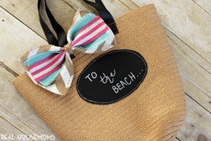 $1 Spot DIY Personalized Beach Tote. Straw sack with black handles, large chalkboard label on the front that reads "To the beach" an oversized burlap bow in Spring or Summer colors.