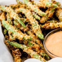 square image of air fryer green bean fries in abasket next to a bowl of sauce