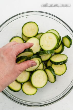 hand tossing zucchini slices in olive oil in a glass bowl