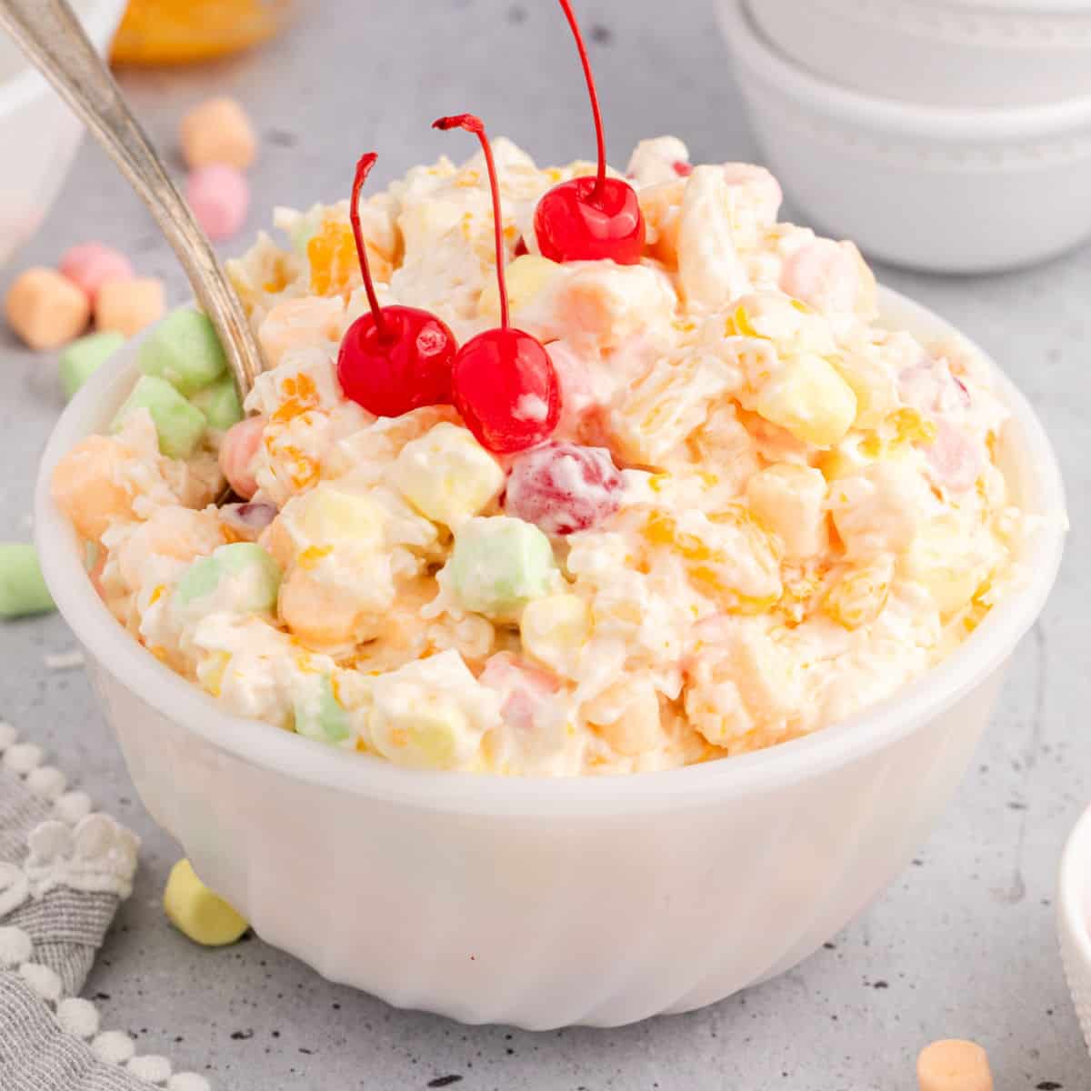 square image of ambrosia salad topped with cherries in a serving bowl with a spoon