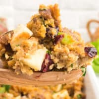 image of a serving spoonful of apple & cranberry cornbread dressing over the baking dish with the title of the post on the top in cranberry and black lettering