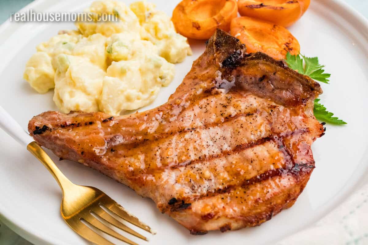 apricot grilled pork chops on a plate with potato salad, grilled apricot halves, and a fork