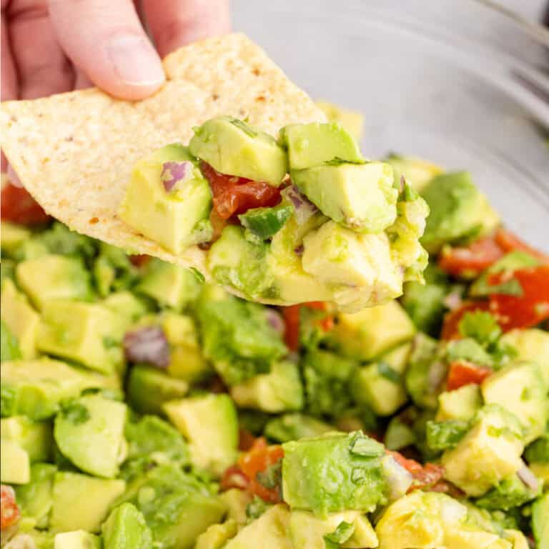 square image of a hand holding a tortilla chip with avocado salsa on it