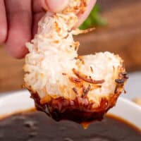 image of a hand dipping a baked coconut shrimp in the garlic plum dipping sauce with the title on top with black and pink lettering