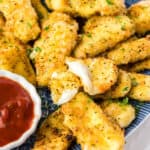 square image of baked mozzarella sticks on a plate next to a bowl of marinara with one broken open to show gooey center