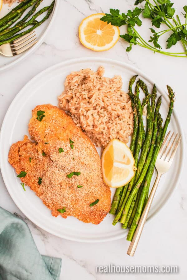baked parmesan tilapia on a plate with asparagus, brown rice, and a lemon wedge