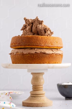 two yellow cake tiers with chocolate frosting between them and big pile of frosting on top ready to be spread