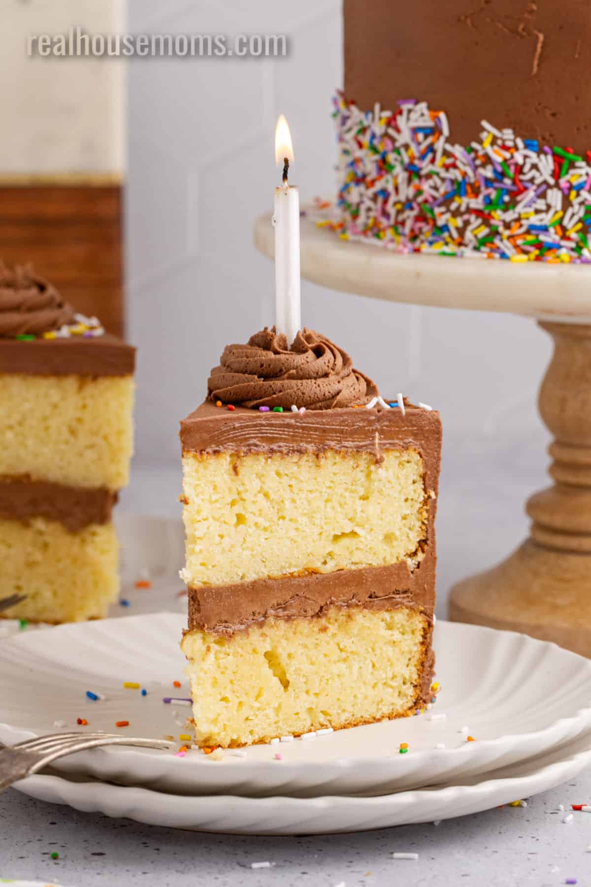 slice of birthday cake with a lit candle on a small plate next to the rest of the cake