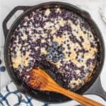 square image of blueberry dump cake in a the skillet with a wooden spoon resting where a portion has been taken out