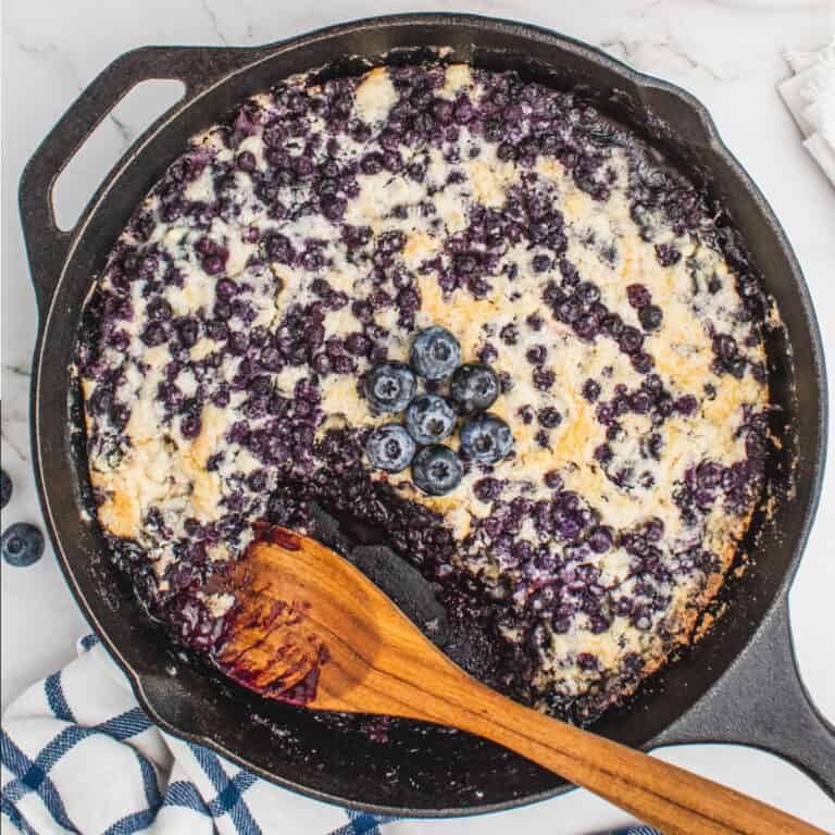 square image of blueberry dump cake in a the skillet with a wooden spoon resting where a portion has been taken out