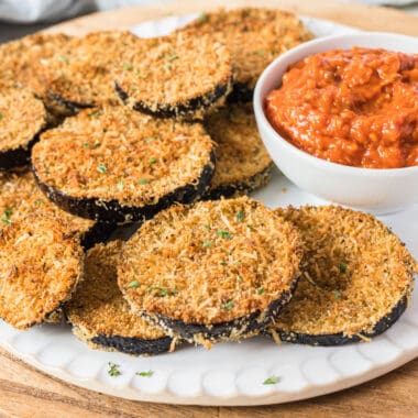 square image of breaded eggplant slices on a plate with a bowl of marinara