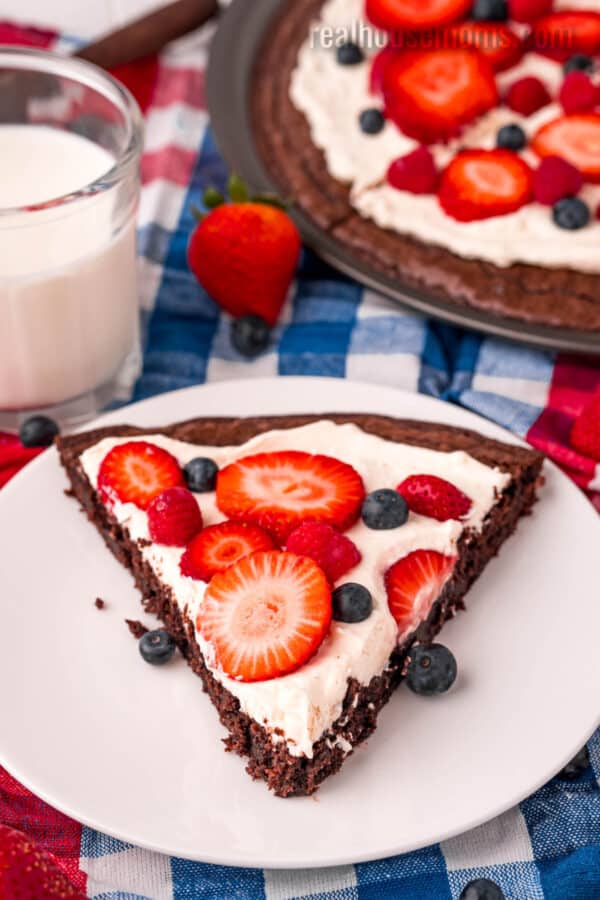 slice of brownie fruit pizza on a plate in front of the pizza and a glass of milk