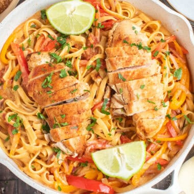 square image of sliced cajun chicken breasts resting on sun-dried tomato alfredo fettuccine in a skillet with lime halves