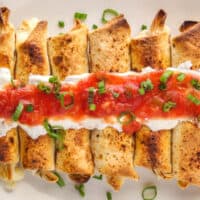 square image of cheesy baked beef taquitos on a platter with sour cream, salsa, and sliced green onion on top