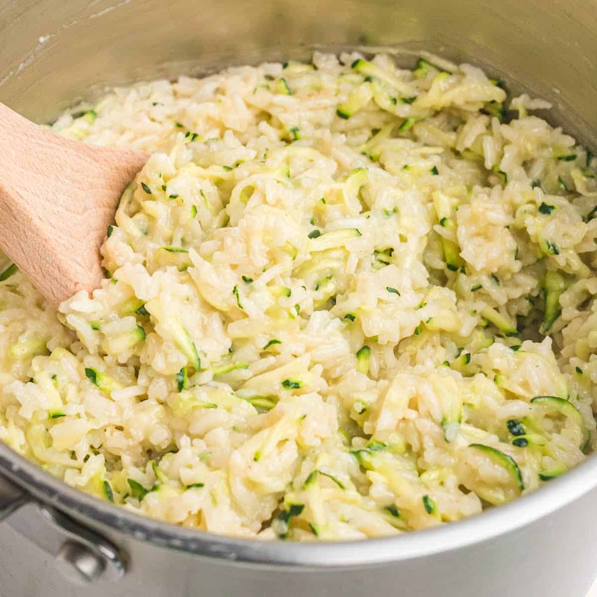 square image of cheesy zucchini rice in a saucepan with a wooden spoon