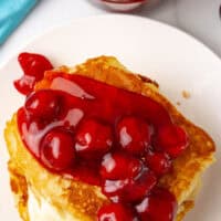 looking down at cherry cheesecake pancakes on a plate next to a jar of cherry pie filling with the title of the post on top of the image in red and black lettering