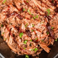 cherry cola pulled pork in a slow cooker topped with a drizzle of bbq sauce and chopped parsley with recipe name at the bottom