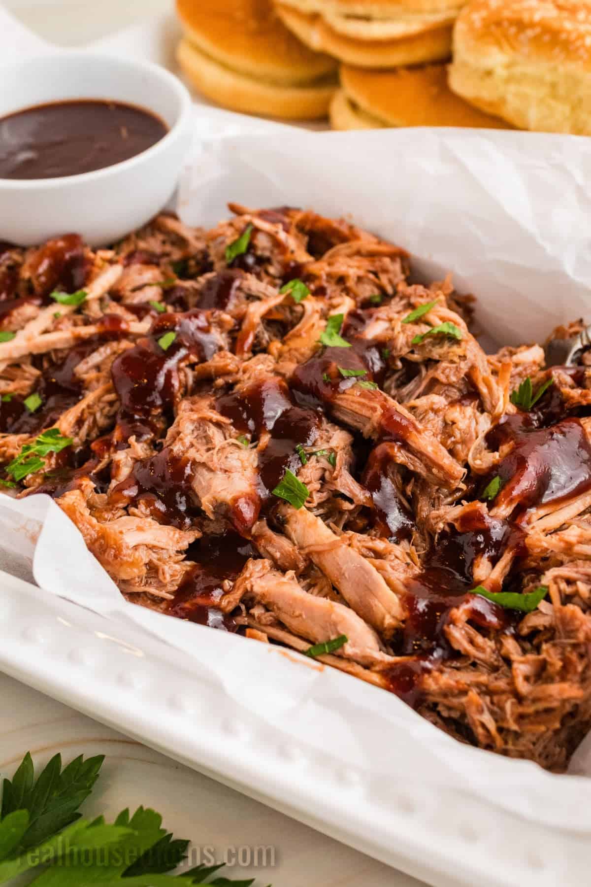 cherry cola pulled pork drizzled with bbq sauce in a serving bowl next to a bowl of sauce and buns