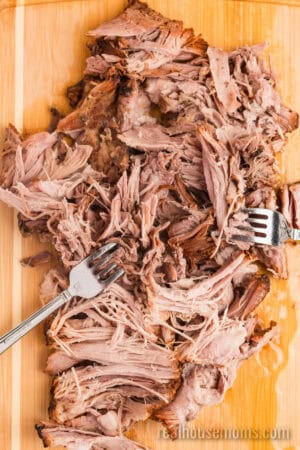 two forks shredding cherry cola pork shoulder on a cutting board