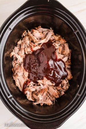 shredded cherry cola pork in a slow cooker with bbq sauce poured over top