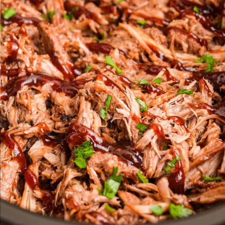 square image of cherry cola pulled pork topped with a drizzle of bbq sauce and chopped parsley in a slow cooker