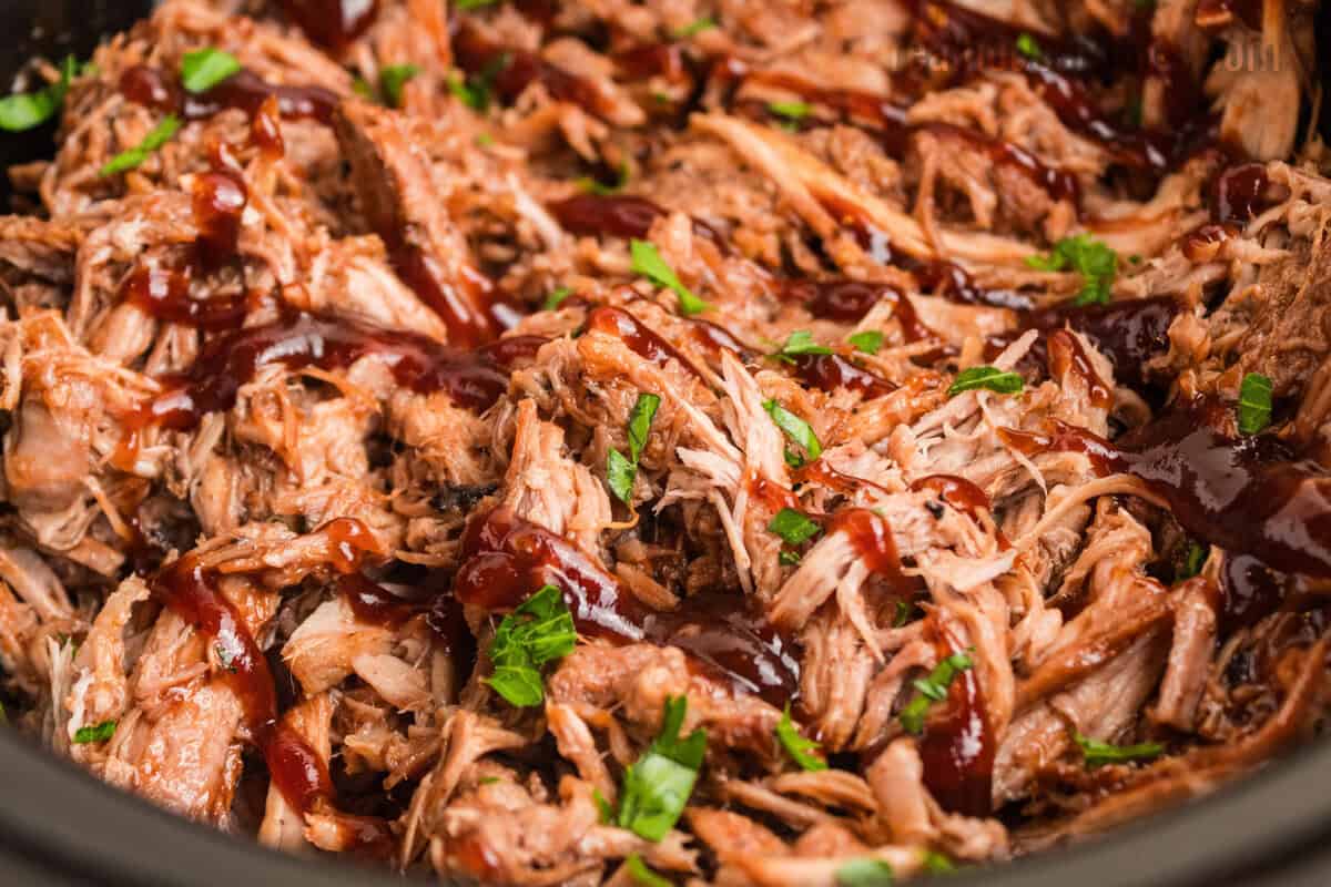 cherry cola pulled pork topped with chopped parsley and bbq sauce drizzle in a slow cooker