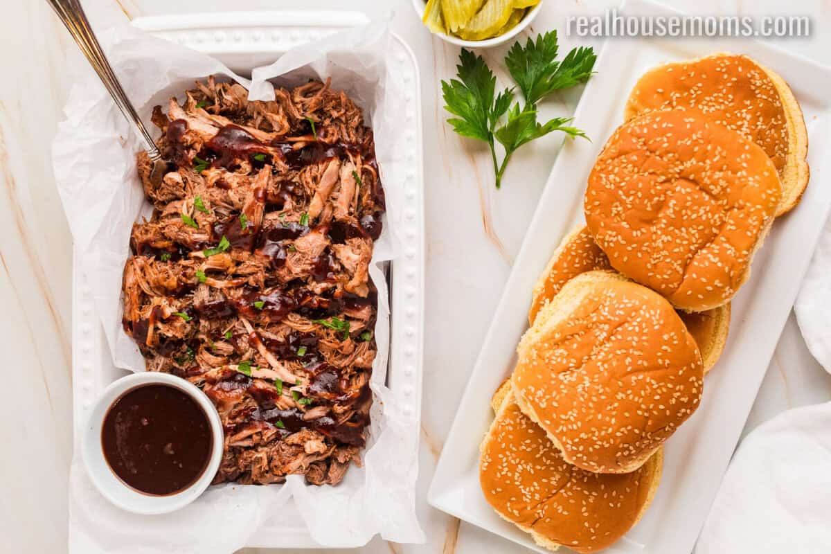 cherry cola pulled pork and a bowl of bbq sauce in a tray next to sandwich buns with sesame seeds
