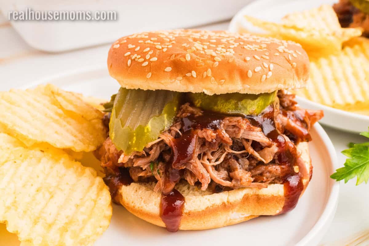 cherry cola pulled pork sandwich with bbq sauce and pickles on a plate with potato chips