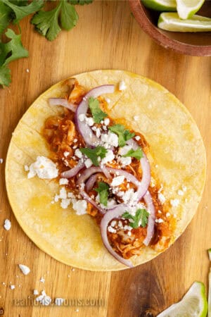 corn tortilla topped with chicken tinga, cotija, cilantro, and sliced red onion on a cutting board