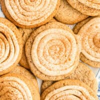 cinnamon roll sugar cookies piled on a plate next to some cinnamon sticks with the title of the post on top of the image in pink and black lettering