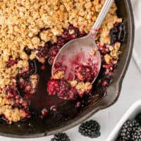 serving spoon in a skillet of blackberry crumble with recipe name at the bottom