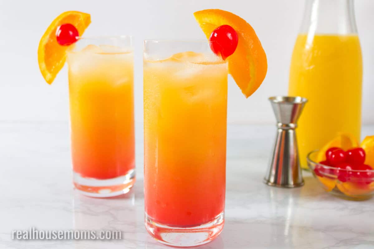 two tequila sunrise cocktails next to a carafe of orange juice