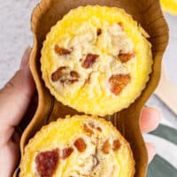 close up hand holding two copycat starbucks bacon & gruyere egg bites in a brown paper holder with the title of the post on top of the image in green and black lettering