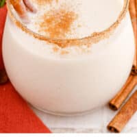 coquito in a cinnamon-sugar rimmed glass with a cinnamon stick with recipe name at the bottom