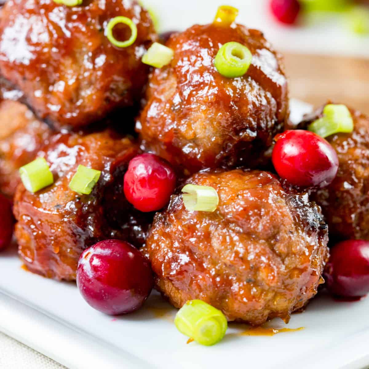 square image of cranberry bbq meatballs on a serving plate with fresh cranberries and sliced green onion