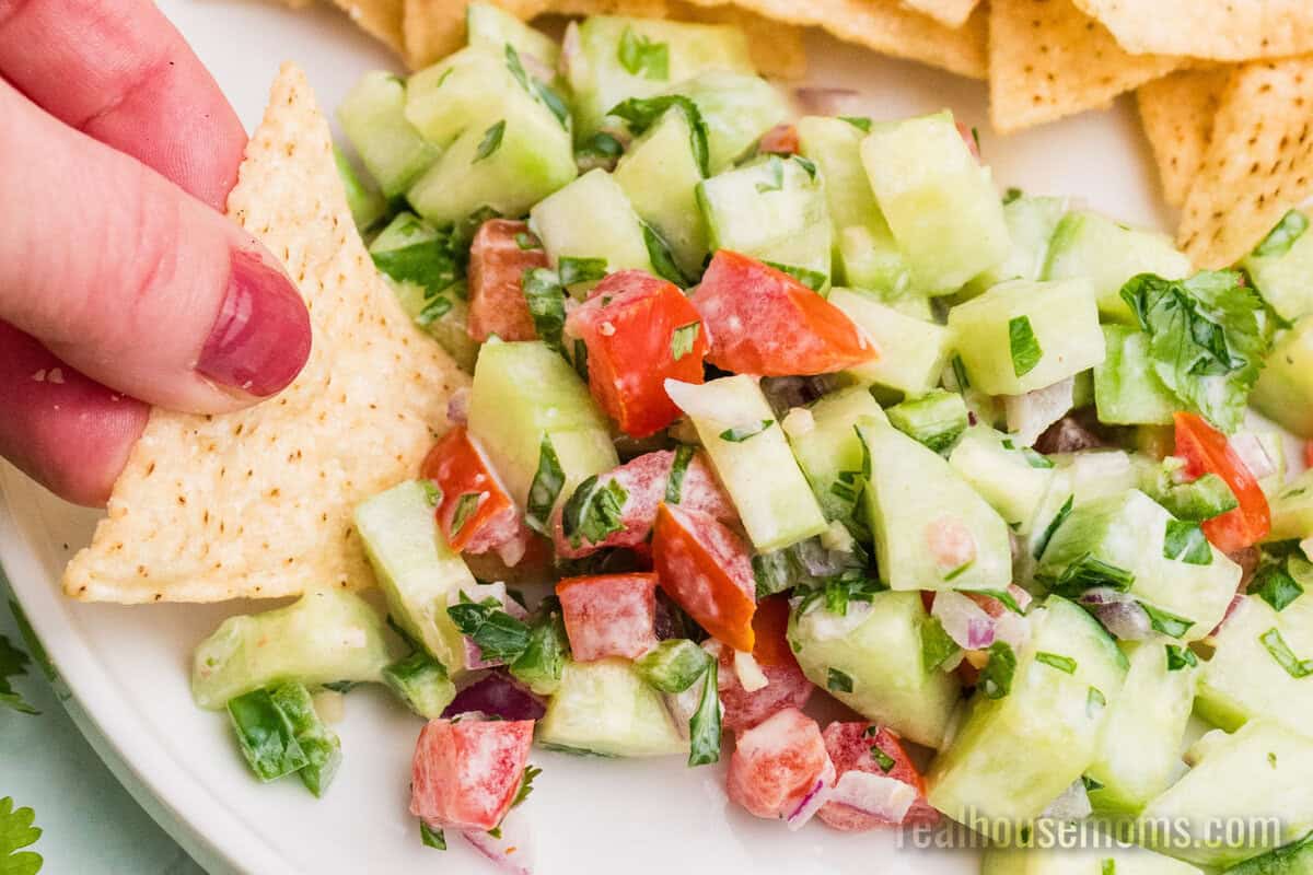 hand dipping a tortilla chip into creamy cucumber salsa on a plate next to more chips
