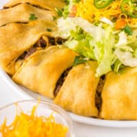 taco crescent roll ring on a serving plate with pico de gallo and shredded cheese with recipe name at bottom