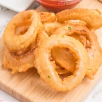 crispy onion rings piled on a wooden board next to a bowl of ketchup with recipe name at the bottom