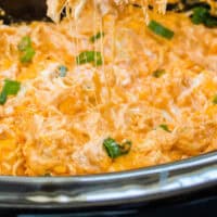 tortilla chip dipped in Crock Pot Buffalo Chicken Dip being lifted by a hand with recipe name at the bottom