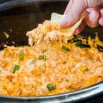 square image of a tortilla chip dipped in Crock Pot Buffalo Chicken Dip being lifted by a hand