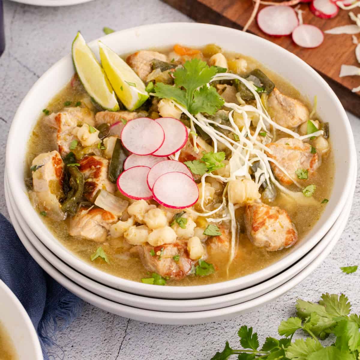 square image of crock pot pozole in a bowl topped with cabbage, radish slices, cilantro, and lime wedges