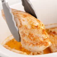 square image of tongs holding a crockpot chicken breast over the slow cooker