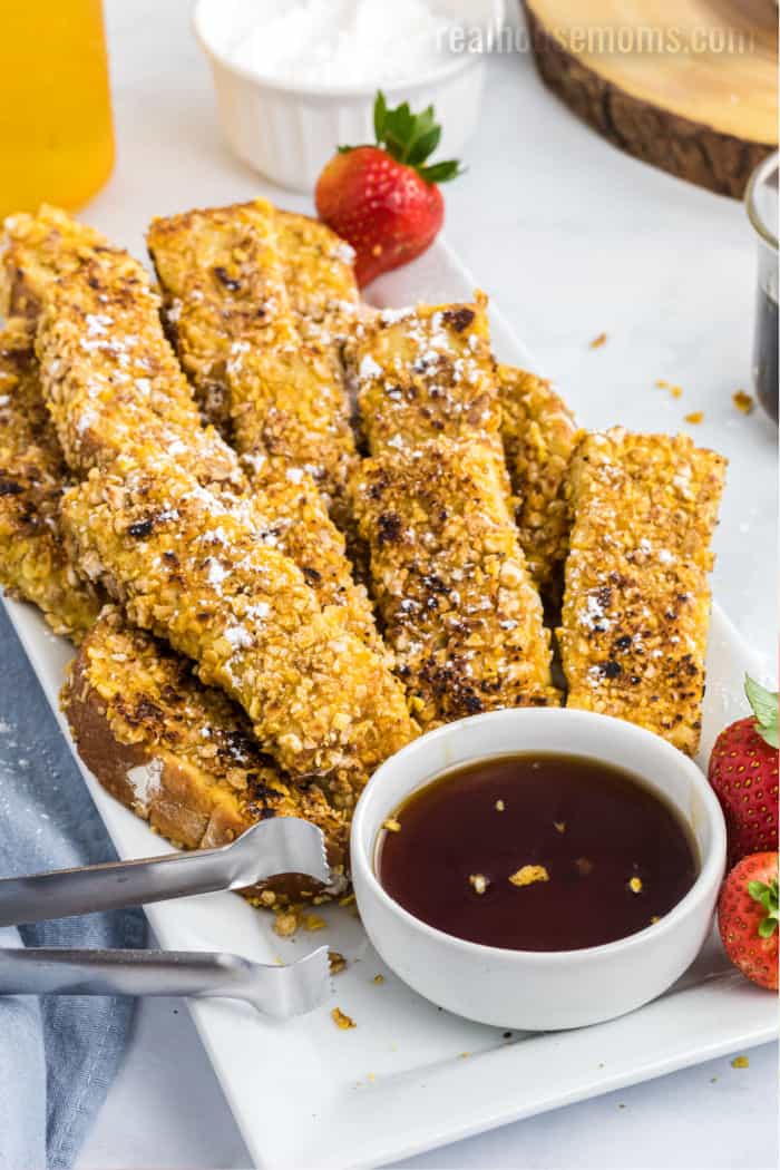 crunchy french toast sticks on a platter with tongs and a bowl of syrup for dipping