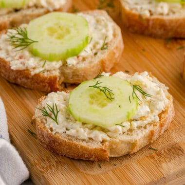 square image of cucumber bruschetta topped with fresh dill on a wooden board