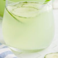 glass of cucumber punch in front of the pitcher with recipe name at the bottom