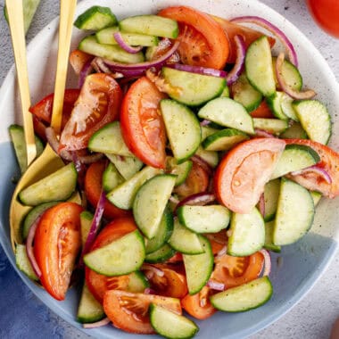 square image of cucumber tomato salad in a serving bowl with gold spoons resting on one side